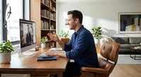 Middle-aged Caucasian businessman in blue sweater having video call at home office desk with computer, smiling during remote work meeting in modern living space.