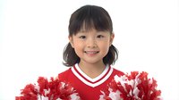 Young Asian girl with pigtails wearing red and white cheerleader uniform, holding pom-poms and smiling brightly against white background.