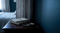 Stacked books and eyeglasses on wooden nightstand in modern bedroom with blue walls and curtains, suitable for education and lifestyle content.