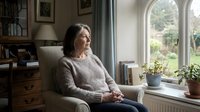 Middle-aged woman with long dark hair and fair complexion, wearing a cozy knit sweater, sitting thoughtfully in an armchair by a bright window in a home library setting.