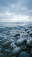 Serene ocean waves washing over smooth pebbles on rocky shore under dramatic cloudy sky creating peaceful coastal atmosphere for meditation and relaxation.