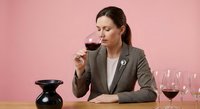 Professional Caucasian businesswoman in gray suit tasting red wine at elegant table with glasses and decanter on pink background for sommelier training.