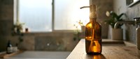 Amber glass pump bottle with golden sunlight on wooden bathroom counter near bathtub, creating warm spa atmosphere for natural skincare routine.