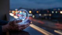 Hands holding glowing digital shield icon with cybersecurity protection symbol against blurred city lights background at dusk for technology concepts.