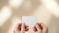 Female hands holding blank white business card against soft beige background with natural lighting for mockup and branding design projects.