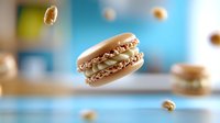 Floating beige macaron cookie with creamy filling against blurred blue background, dynamic food photography with levitating crumbs creating sense of movement.