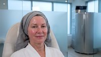 Smiling mature Caucasian female healthcare professional in medical hijab and white coat sitting in modern hospital or clinic office environment.