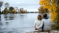 Couple sitting on riverbank enjoying autumn view with vibrant yellow foliage and calm water reflecting cloudy sky.