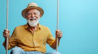 Happy senior Caucasian man with white beard wearing straw hat and yellow shirt sitting on rope swing against blue background. Joyful retirement lifestyle concept.