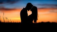 Romantic silhouette of young couple touching foreheads at sunset with dramatic orange sky and wild grass, creating intimate moment in nature.
