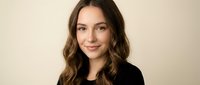 Young professional Caucasian woman with wavy brown hair smiling confidently in black blazer against neutral beige background for business portrait.