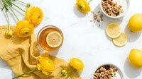 Herbal tea with lemon slices and yellow ranunculus flowers on white background. Healthy morning beverage with granola and citrus fruits flat lay composition.