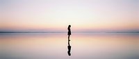 Silhouette of woman walking on reflective wet sand beach at sunset with pink purple sky and calm ocean creating mirror reflection for meditation wellness concepts.