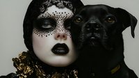 Gothic woman with ornate black lace mask and dark makeup poses with black dog companion in dramatic portrait style for Halloween or fantasy themes.
