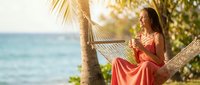 Young woman in coral dress relaxing on hammock with tropical drink at beach resort during golden hour sunset vacation getaway.