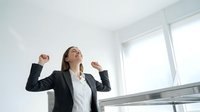 Young professional businesswoman celebrating success with raised arms in modern office environment expressing joy and achievement at workplace.