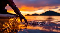 Hand touching water surface creating splash and ripples during dramatic orange sunset over mountain lake, silhouette against vibrant sky reflections.