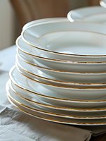 Stack of elegant white porcelain dinner plates with delicate gold trim on white linen tablecloth, showcasing fine dining tableware and luxury table setting.