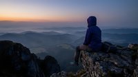 Person in dark blue jacket sitting on rocky mountain peak at sunrise, overlooking misty valley landscape below.