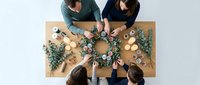 Four people collaborating on creative Christmas wreath making workshop at wooden table with eucalyptus branches and craft supplies for holiday decoration project.