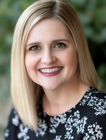 Confident middle-aged Caucasian woman with blonde hair and bright smile wearing floral black dress against blurred green background, professional headshot style portrait.