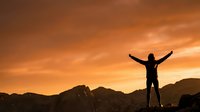 Person celebrating success on mountain peak at golden sunset with arms raised in triumph against dramatic orange sky and mountain silhouette landscape.