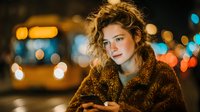 Young woman with curly hair using smartphone at night in urban setting with bokeh lights. Evening lifestyle portrait for social media and technology concepts.