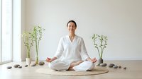 Young Asian woman in white clothing practicing meditation in lotus position on woven mat surrounded by bamboo plants and stones in minimalist room.