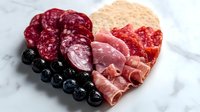 Heart-shaped charcuterie board with assorted cured meats, salami, prosciutto, crackers and fresh blueberries on marble surface for romantic dining.