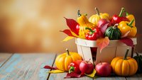 Autumn harvest basket filled with colorful pumpkins, gourds and fall leaves on rustic wooden table with warm golden background for seasonal decoration.