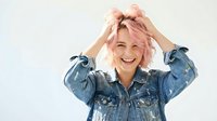Young woman with short pink hair wearing denim jacket, hands in hair, smiling at camera against white background for fashion and lifestyle content.