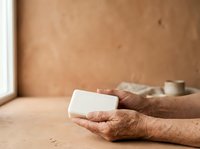 Senior adult with dark skin tone holding white soap bar on neutral beige surface, promoting natural skincare, hygiene products, wellness routines, and dermatological care for mature skin.