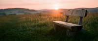 Wooden bench sitting alone in a grassy meadow during golden hour sunset, with rolling hills and warm sky in background for peaceful outdoor concepts.