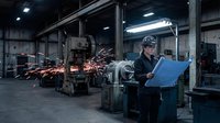 Worker in dark uniform and hard hat reviewing blue clipboard in industrial factory with welding sparks flying from machinery in background.