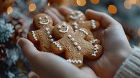 Freshly baked gingerbread men cookies with white icing decoration held in hands wearing cozy sweater against blurred holiday lights background.