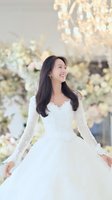 Young Asian bride in elegant white wedding dress with lace sleeves smiling joyfully at luxury wedding venue with floral decorations and chandelier lighting.