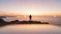 Silhouetted figure standing on rocky outcrop overlooking misty coastal landscape at sunrise, conveying solitude, contemplation, and adventure for travel and lifestyle content.