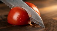 Cherry tomato being sliced with sharp knife on wooden surface, showing fresh produce preparation for cooking and culinary use.