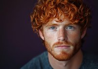Young man with bright red curly hair, ginger beard, and striking blue eyes against dark background. Natural freckles and genuine expression create authentic portrait.