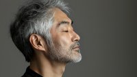 Profile of mature Asian man with gray hair and beard against dark background, eyes closed in serene expression. Minimalist portrait highlighting natural aging and wisdom.