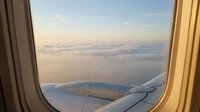 Airplane window view of aircraft wing flying above clouds at sunrise or sunset with golden light illuminating the sky, suitable for travel and aviation concepts.