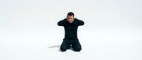 Young man in black clothing kneeling on white background covering ears with hands in distress or overwhelm gesture isolated for design use.
