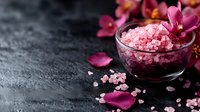 Pink aromatic bath salt in glass bowl with purple orchid petals on dark stone background, spa wellness composition for relaxation and body care.