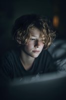 Young boy with curly blonde hair illuminated by screen light in dark room, creating moody atmosphere for digital lifestyle or technology concept.