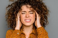 Young woman with curly hair experiencing headache touching temples with pained expression on gray background for health and wellness concepts.
