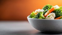 Fresh steamed vegetables with broccoli, cauliflower and carrots in gray ceramic bowl on dark background with warm lighting and copy space.