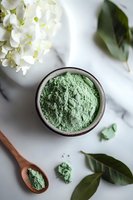 Green clay powder in ceramic bowl with wooden spoon on marble surface, white hydrangea flowers and fresh green leaves creating spa composition.