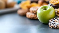 Fresh green apple with assorted cookies and baked treats on dark background, selective focus on fruit. Food photography for healthy versus indulgent choices.