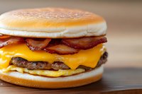 Juicy cheeseburger with crispy bacon strips and fried egg layered between toasted buns on wooden surface, close up view of breakfast sandwich.