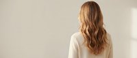 Young blonde woman with long wavy hair wearing white sweater standing with back turned against neutral beige wall in soft natural lighting.
